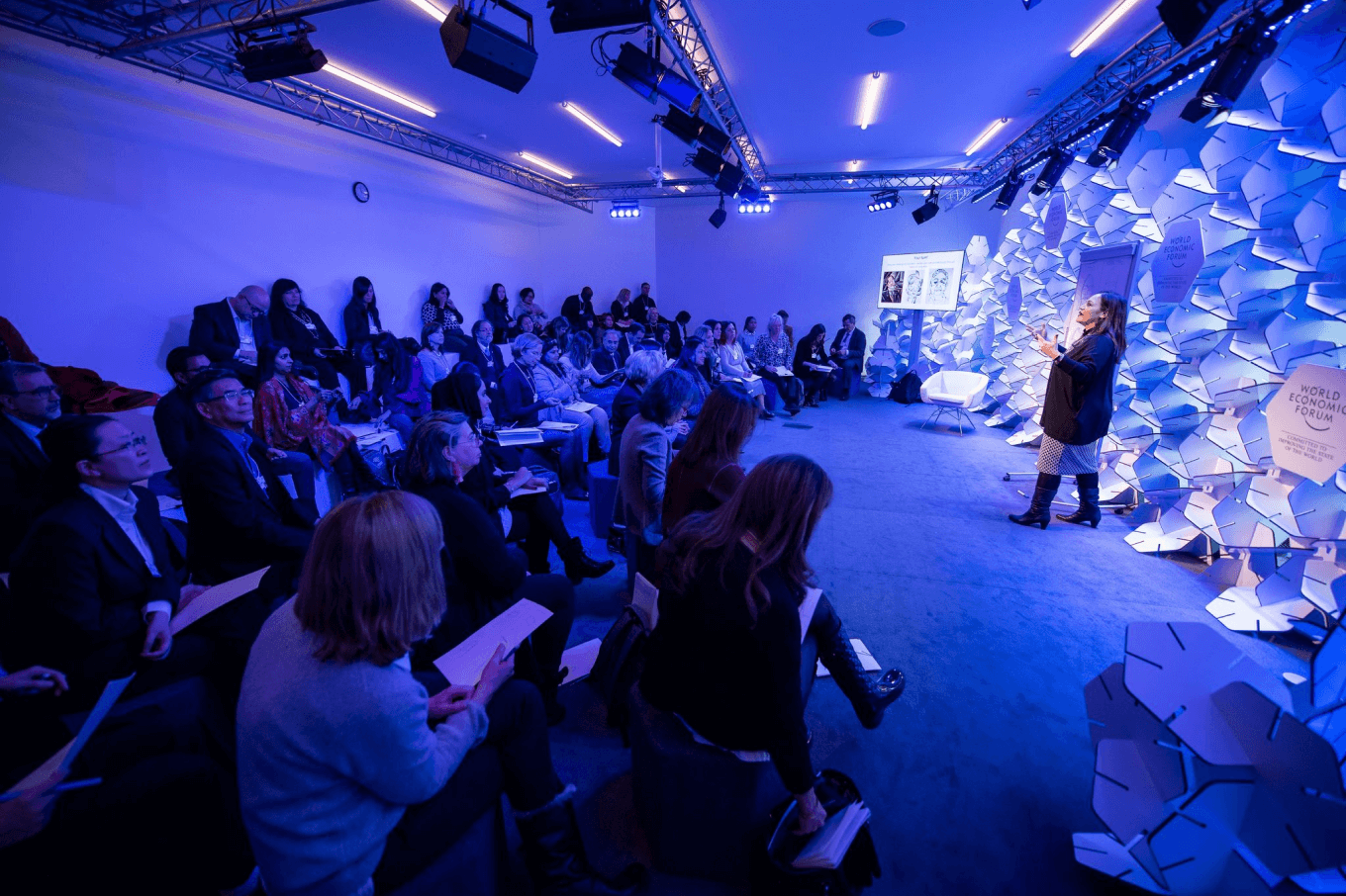 an engaged crowded room listening to a speaker against the backdrop of purple lights